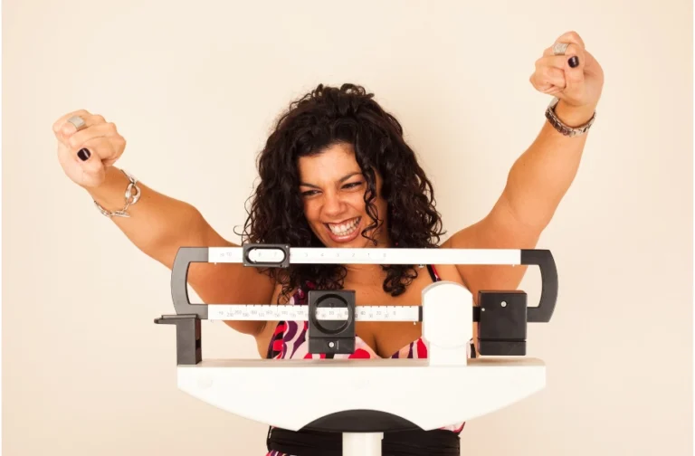 woman with dark curly hair celebrating weight loss on a medical scale