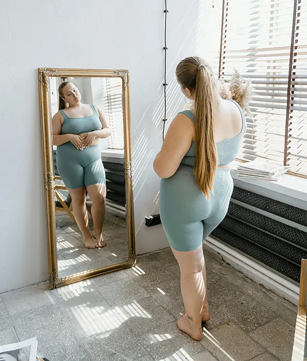 woman in workout attire with a long ponytail looking at herself in a mirror in a neutral colored room filled with natural light